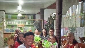Un gruppo di persone sedute a un tavolo di legno in un ristorante, con una ventola a soffitto, piante in vaso e un bancone con diversi oggetti sullo sfondo.
