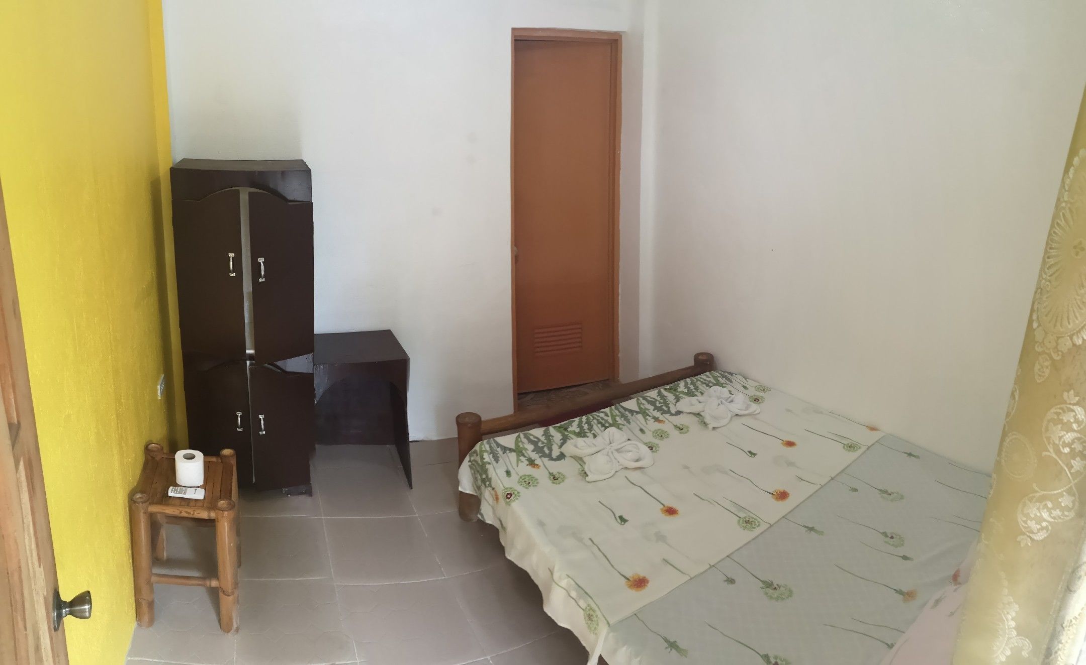 Une petite pièce, peu meublée, avec un lit, une armoire en bois, une petite table sur laquelle se trouve un rouleau de papier toilette, et une porte.