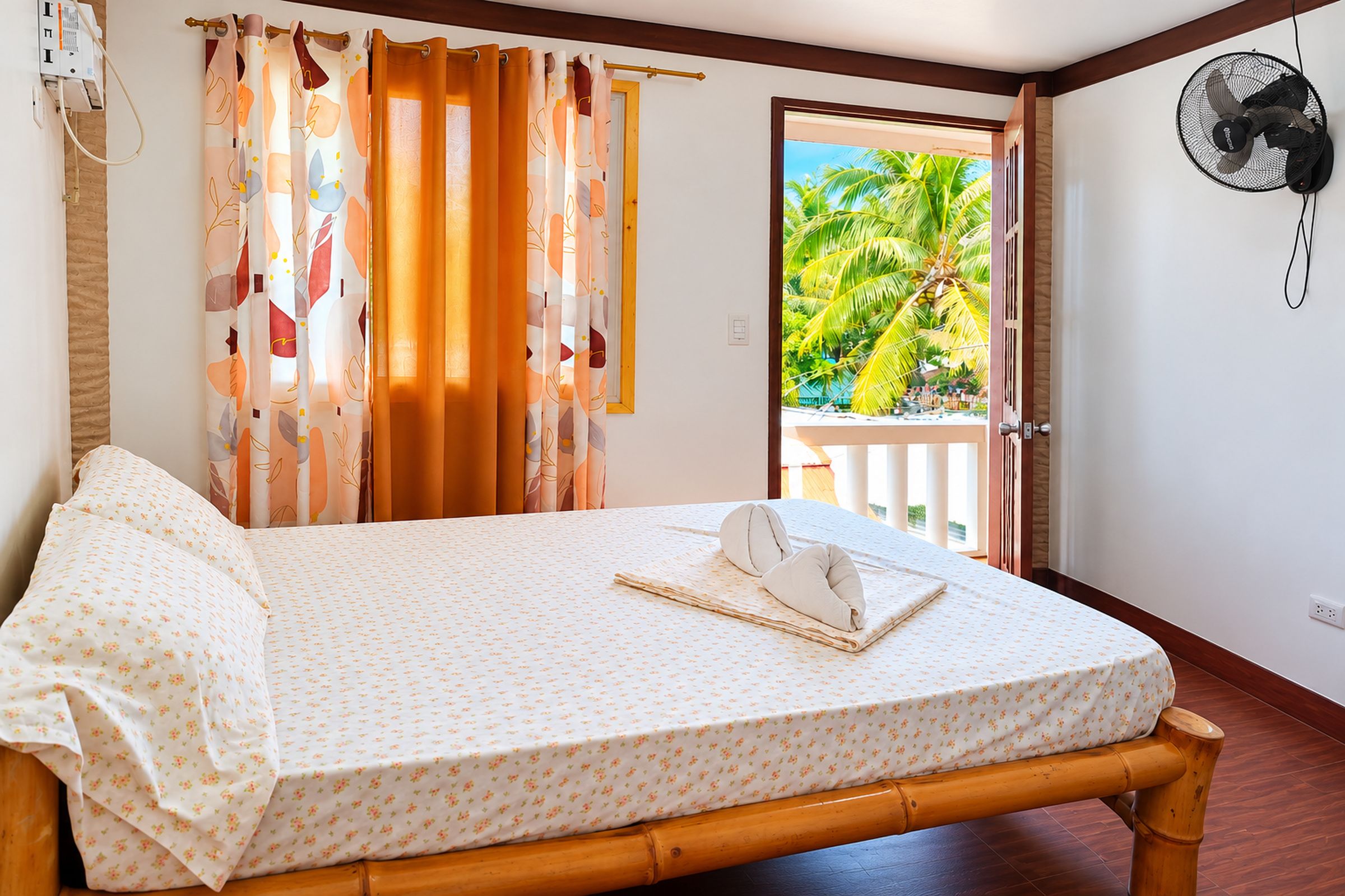 A room with a bed, a fan, and a view of palm trees.