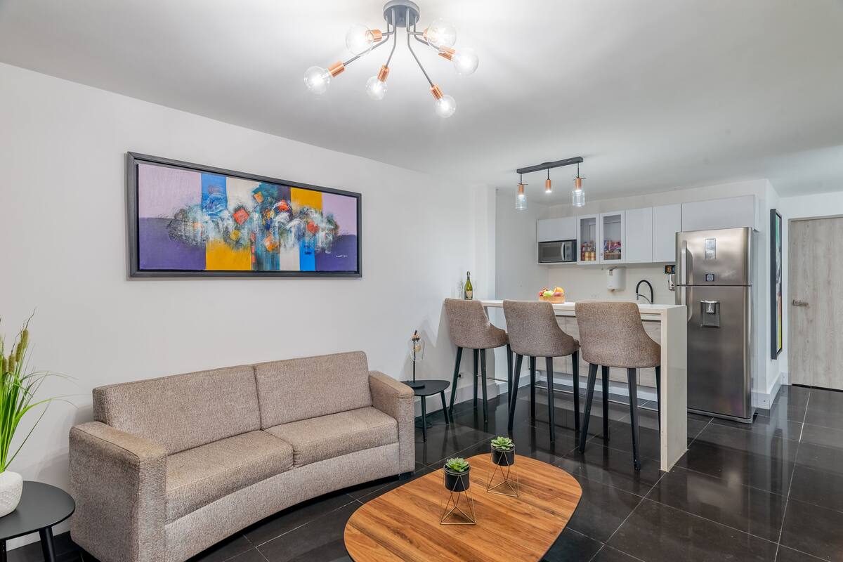 Un salón moderno con un sofá, una mesa de centro y un cuadro. Se ve una cocina con un refrigerador y taburetes de bar al fondo.