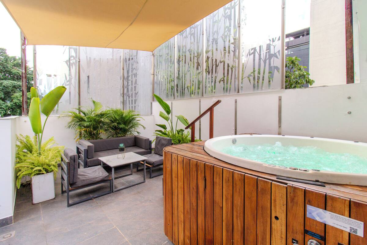 Un jacuzzi con paneles de madera, un sofá, una mesita y plantas en macetas en un patio exterior.