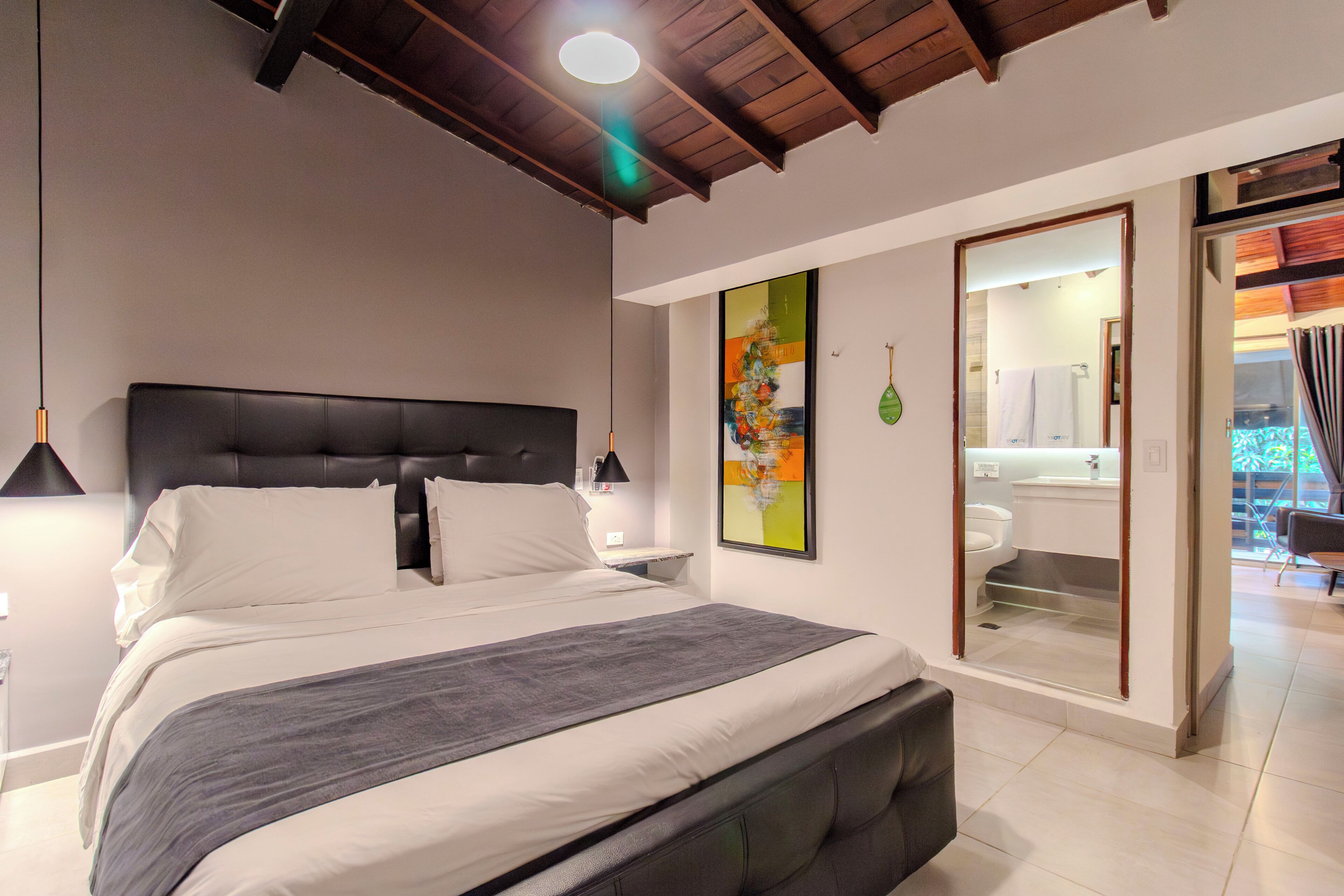 A modern bedroom with a large bed, a nightstand, a wall-mounted lamp, and a bathroom visible through an open door.