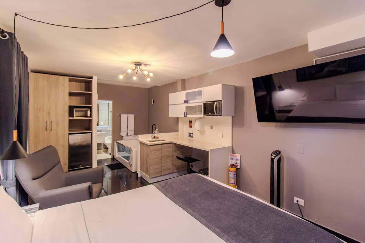 Habitación de hotel moderna con cama, escritorio, silla, kitchenette y televisor montado en la pared.