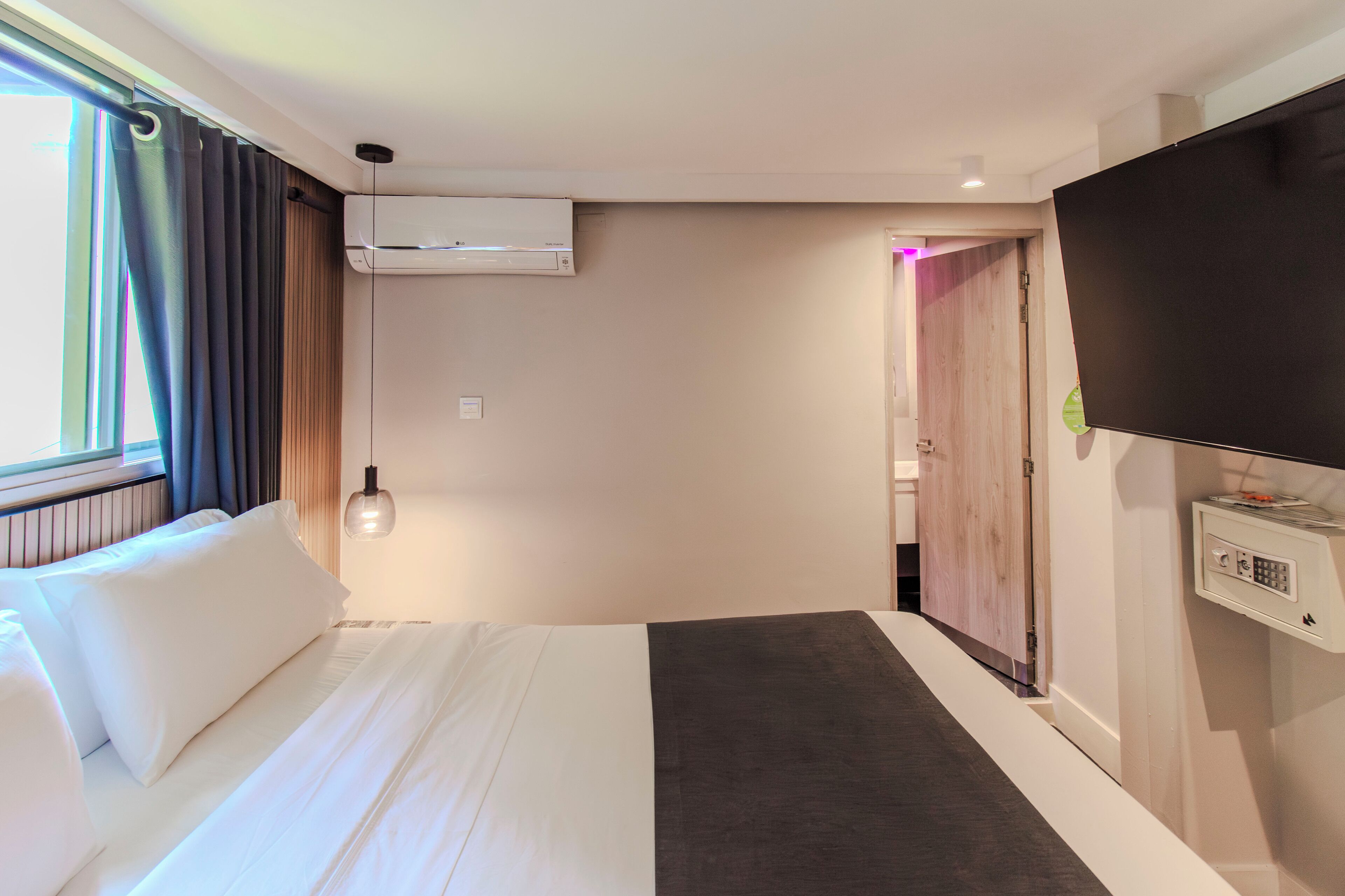 A hotel room with a bed, an air conditioning unit, a television, and a door leading to another room.