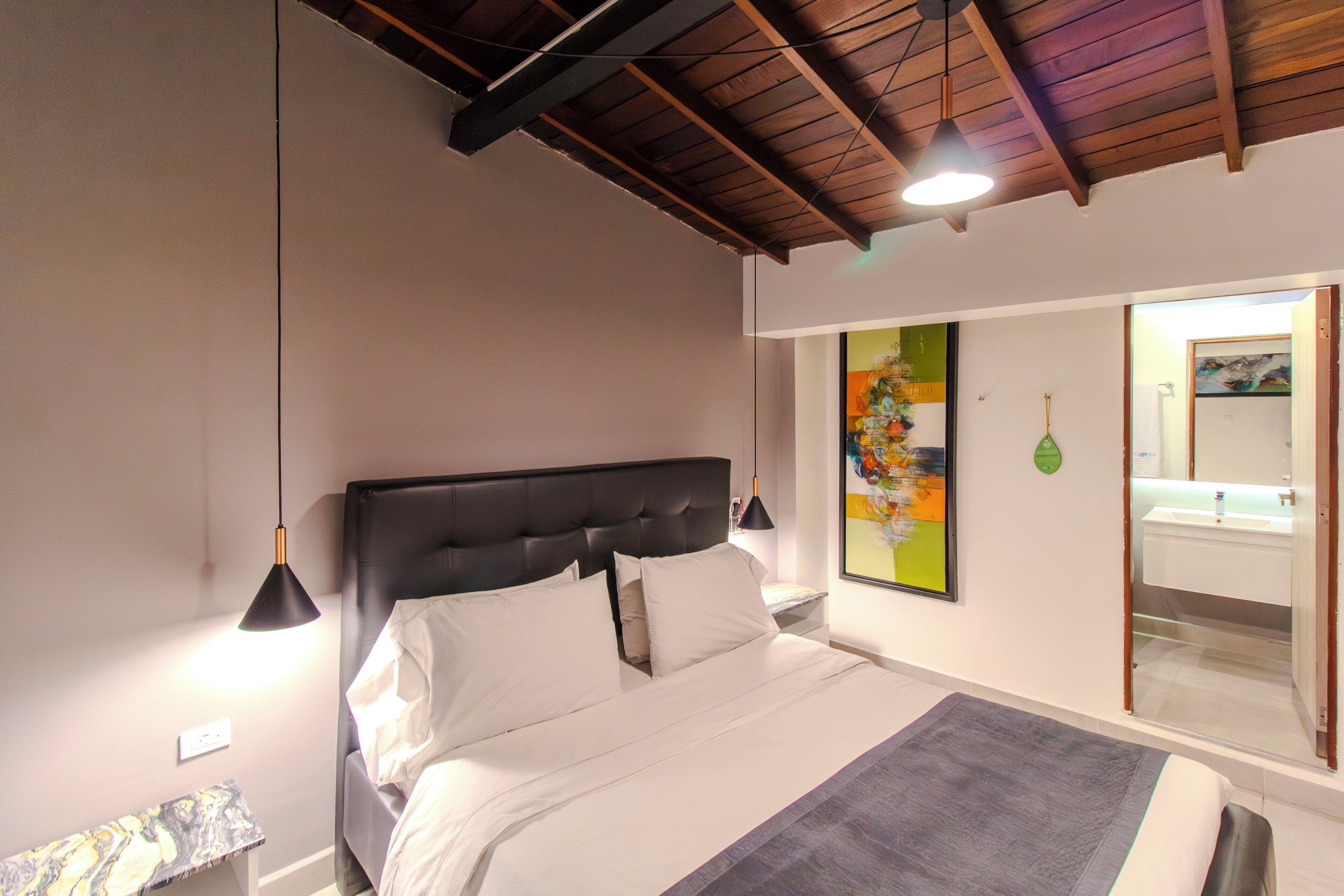 Un dormitorio con cama, lámparas de noche, mesita de noche, un banco, un cuadro y un baño visible a través de una puerta abierta.