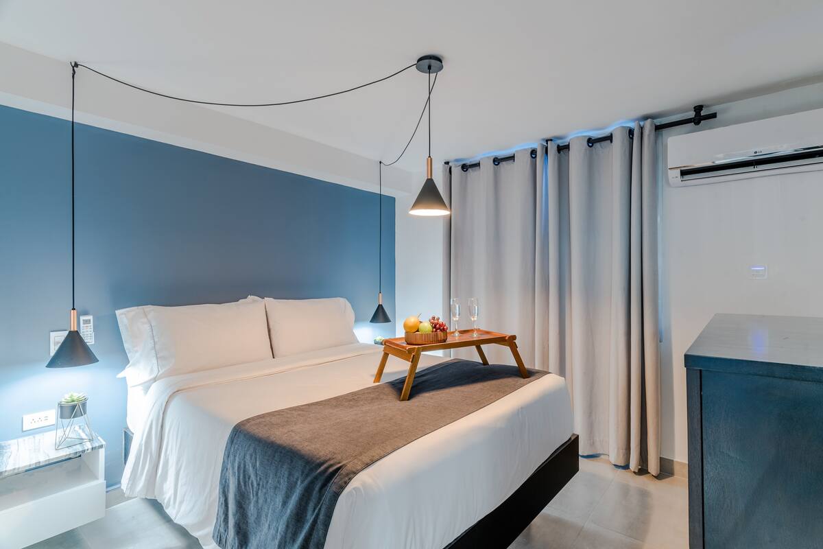 Una habitación de hotel moderna con cama, mesita de noche, lámpara, cortinas y una pequeña mesa con fruta y vasos.