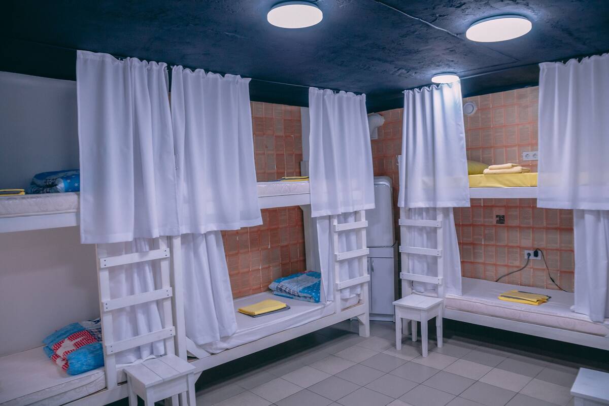 A dormitory room with bunk beds, white curtains, a small table, and a tiled floor.