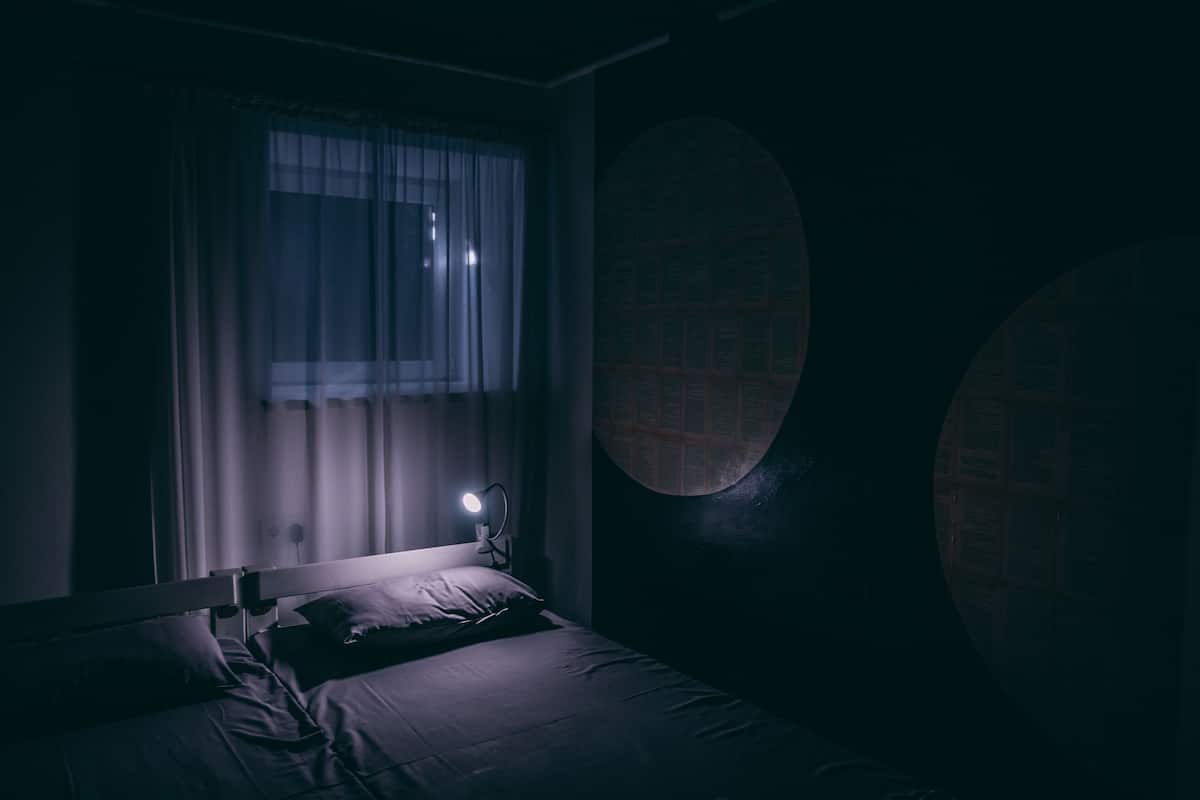 A dimly lit room with a bed, a window with curtains, and a round wall feature.