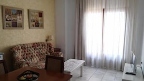 A living room with a floral sofa, a wooden coffee table, and a TV stand. There are two framed pictures on the wall and a window with curtains. - Hotel Al Andalus (Torrox)