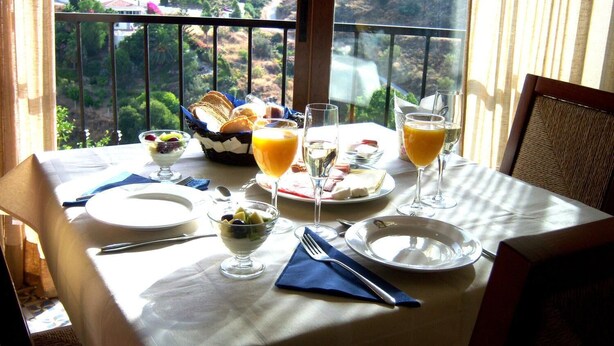 A dining table with breakfast items, including fruit, bread, and drinks, set on a balcony with a scenic view. - Hotel Al Andalus (Torrox)