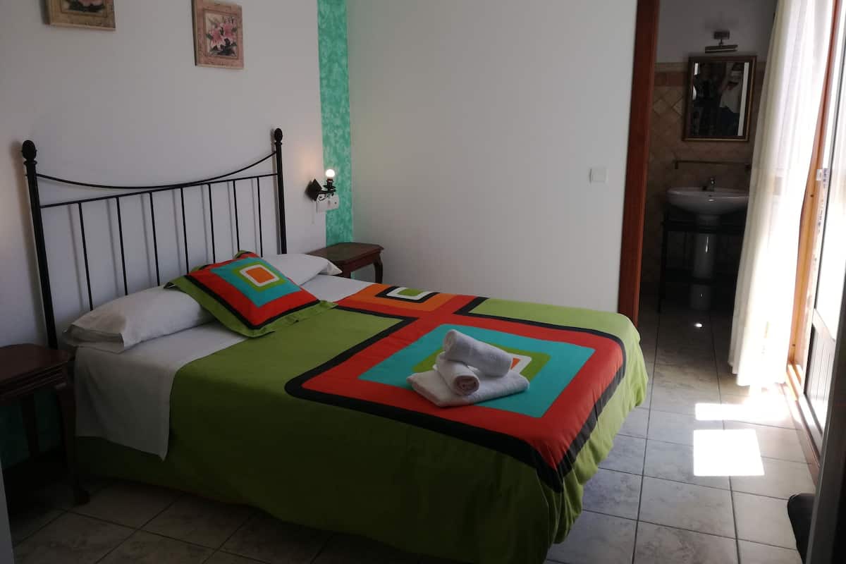 A neatly made bed with a green and red bedspread, two pillows, and a folded towel on the bed. There is a nightstand with a lamp, a wall with two framed pictures, and a bathroom visible through an open door.