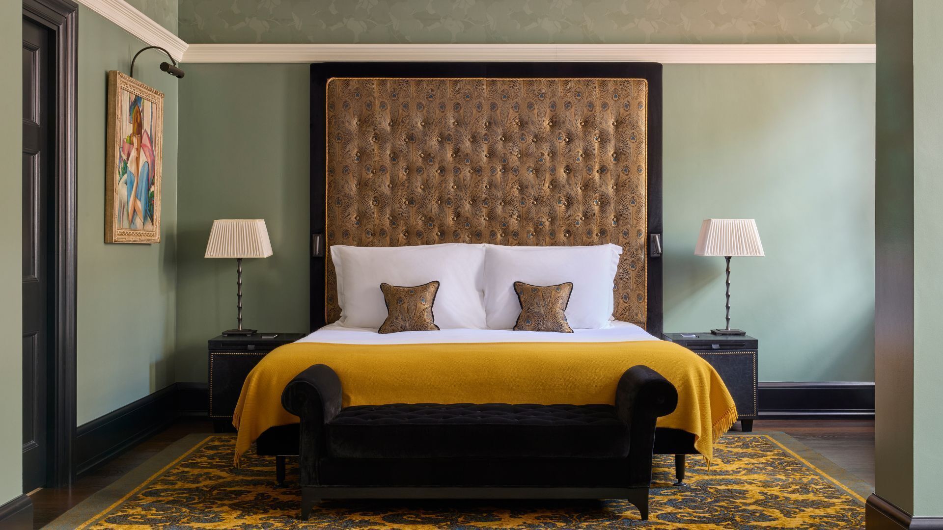 A hotel room with a large bed, two bedside lamps, a bench, and a patterned rug.