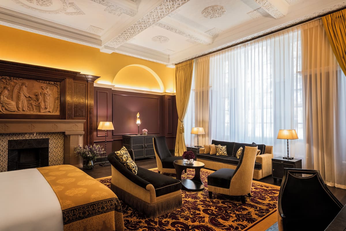 A hotel room with a fireplace, a sofa set, a dining table, and a large window with curtains.