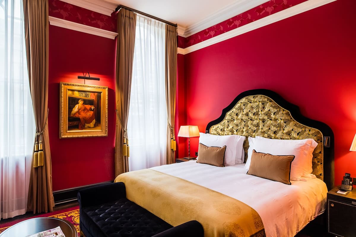 A hotel room with a large bed, a headboard with floral patterns, a painting on the wall, and a desk with a lamp.