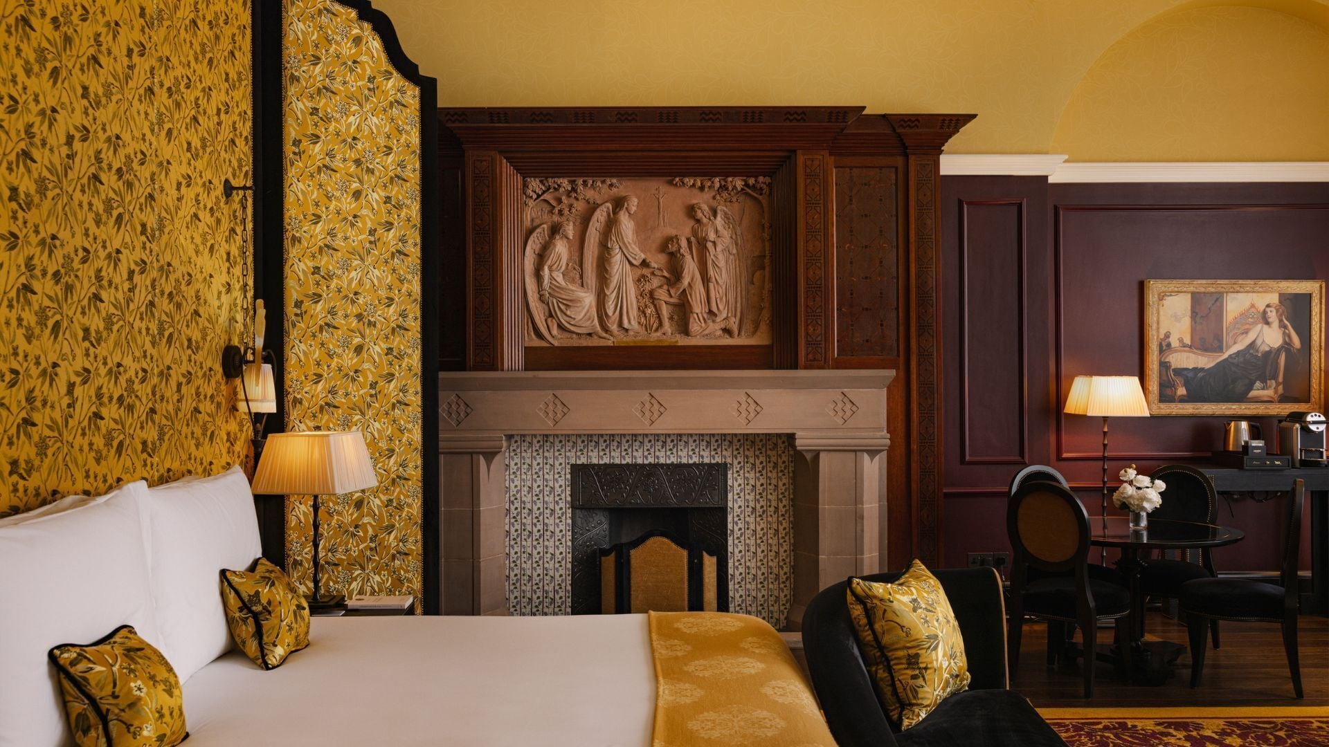 A hotel room with a bed, a fireplace with a relief sculpture, a desk with a lamp, and a painting on the wall.