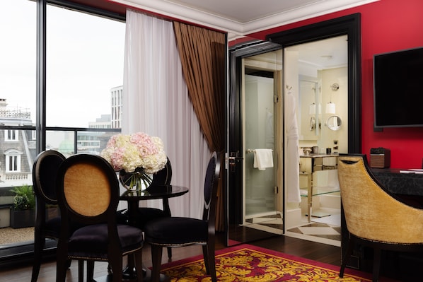 Habitación de hotel con un ventanal grande, un área de comedor con dos sillas, una mesa con un florero y una alfombra roja. Se ve un baño a través de una puerta de cristal.