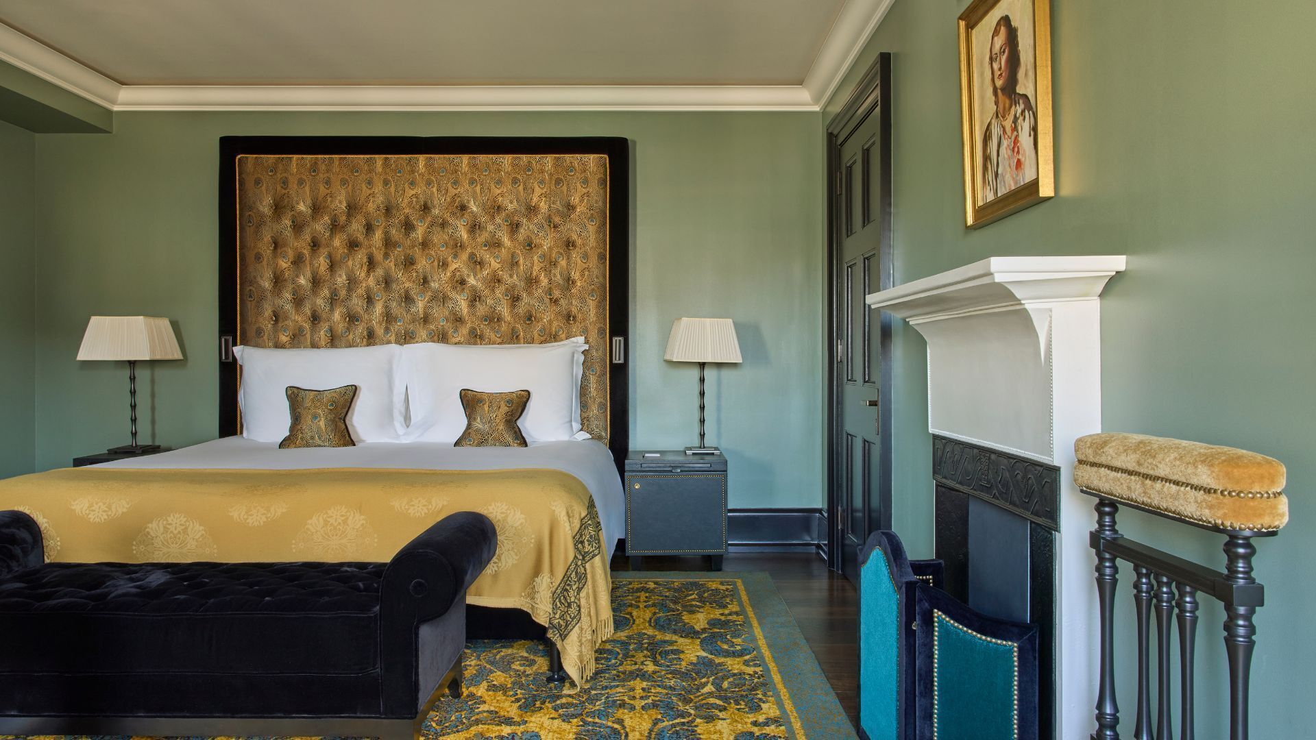 Una habitación de hotel con una cama grande, dos lámparas de noche, un banco tapizado en negro, una alfombra estampada y una chimenea.