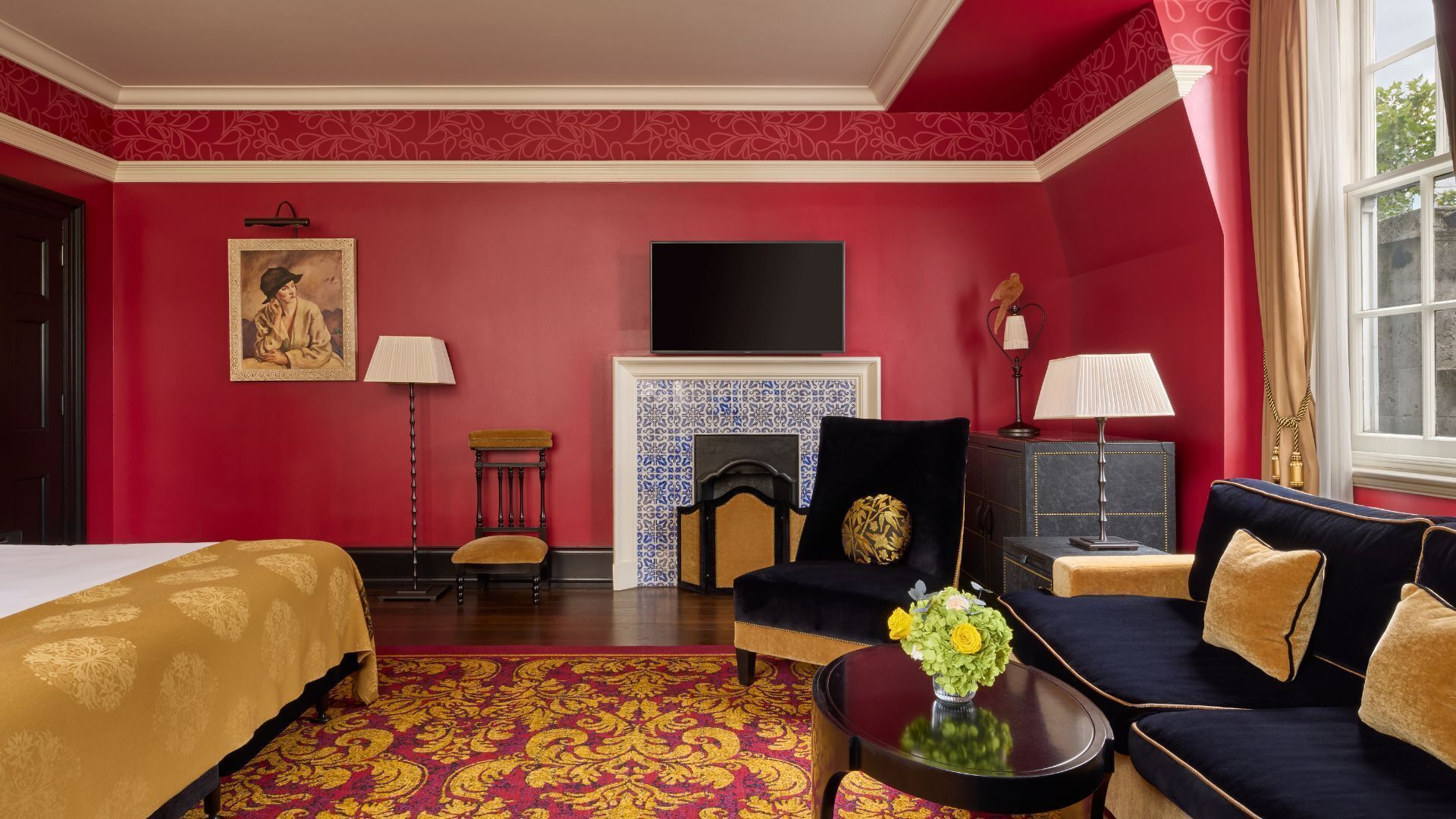 A hotel room with a red wall, a bed with a gold bedspread, a sofa with gold cushions, a fireplace, and a television mounted above it.