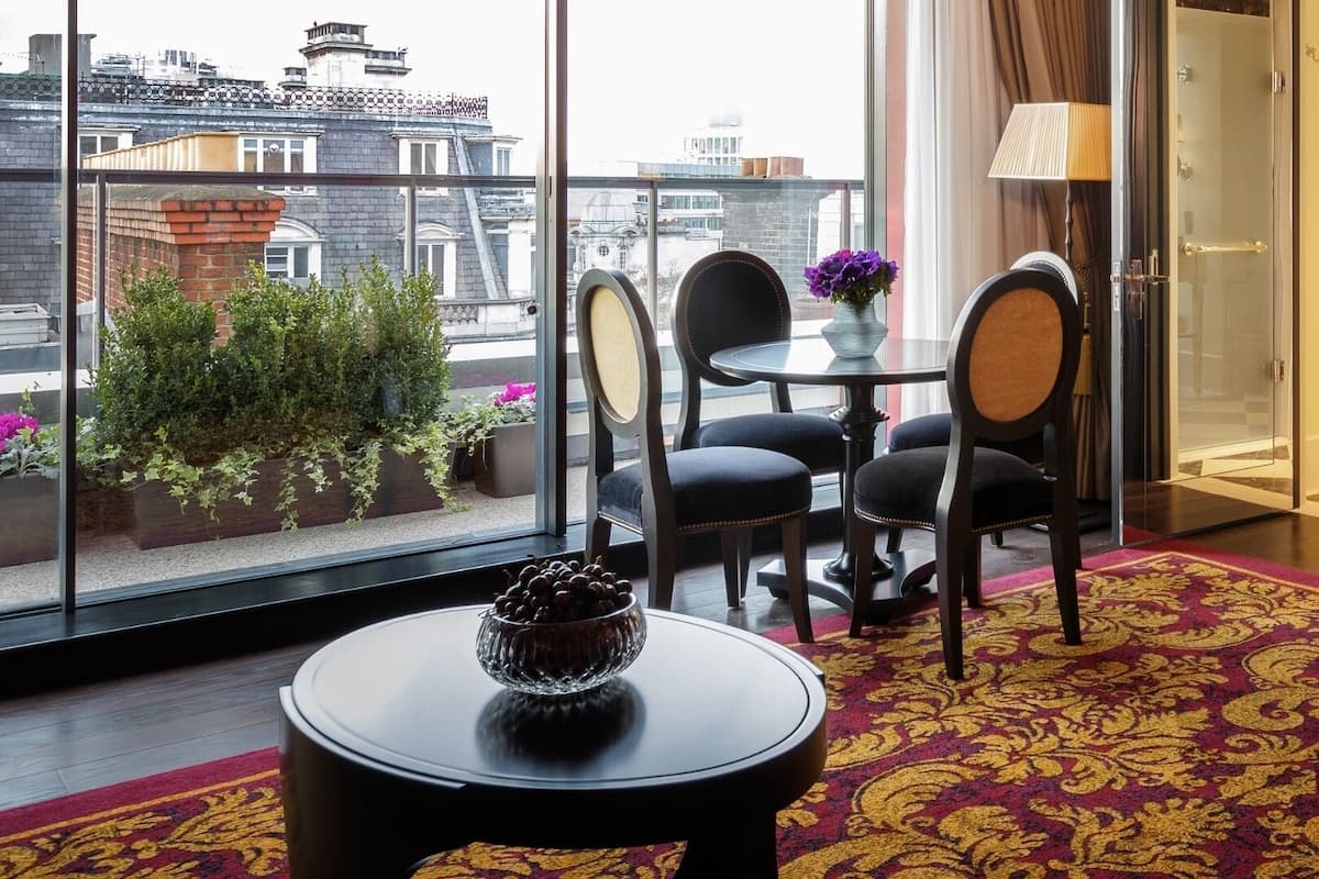 A hotel room with a large window, a round table with chairs, a vase with flowers, and a red carpet.