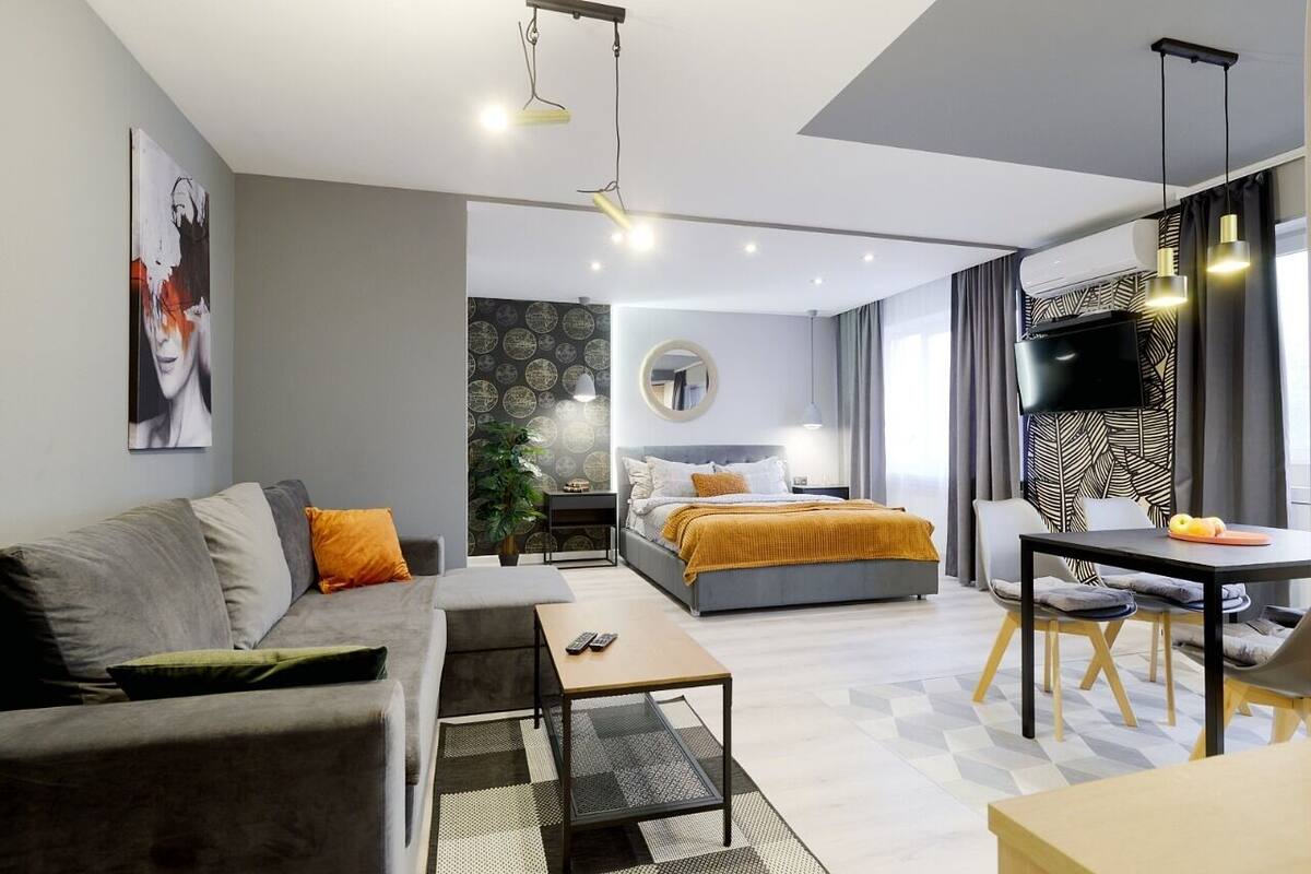 A modern hotel room with a sofa, a bed with a mustard-colored blanket, a dining table, and a TV.