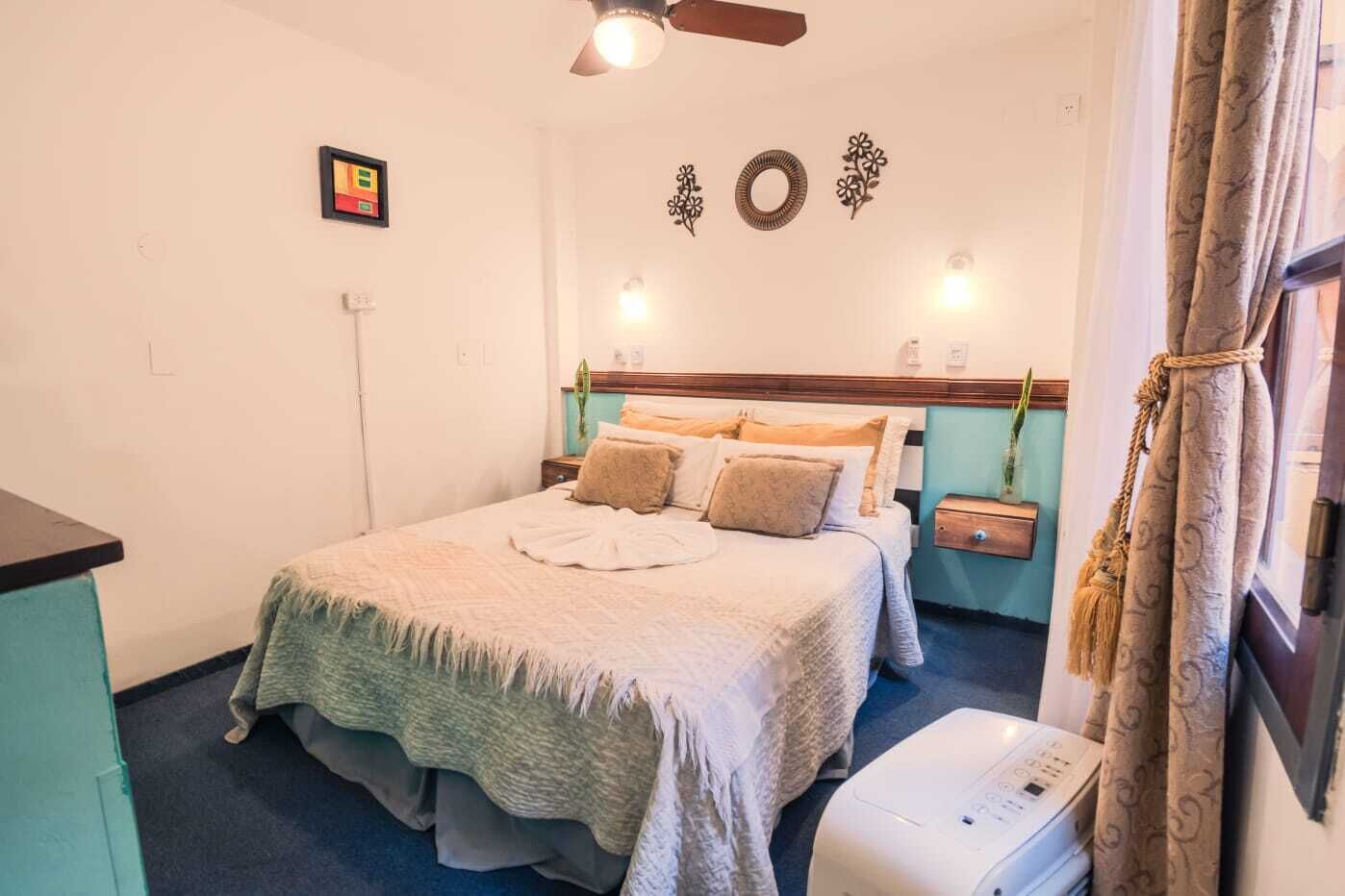 Un dormitorio con cama, mesitas de noche, ventilador de techo, lámpara de pared y ventana con cortinas.