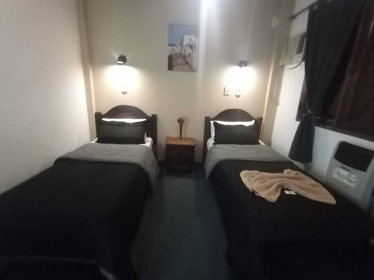 A small, narrow room with two single beds, each with a headboard and a bedside table. There is a small nightstand between the beds, and a wall-mounted shelf above the nightstand. The room has a simple, functional design with minimal decor.