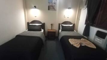 A small, narrow room with two single beds, each with a headboard and a bedside table. There is a small nightstand between the beds, and a wall-mounted shelf above the nightstand. The room has a simple, functional design with minimal decor.
