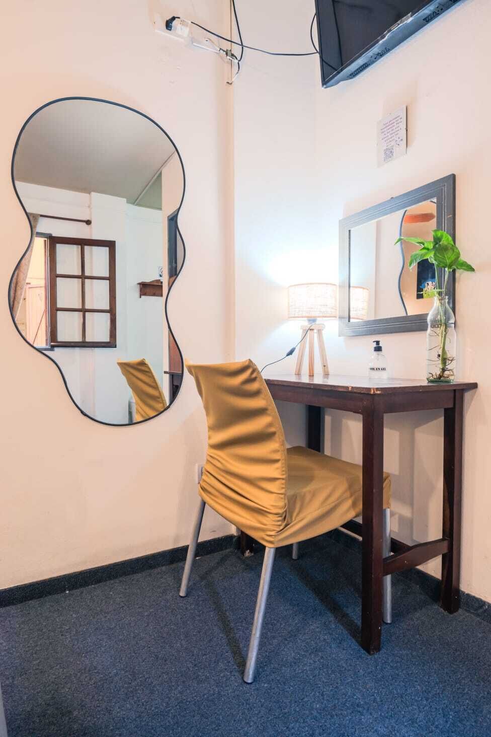 A hotel room with a desk, chair, mirror, lamp, and a plant.