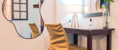 A hotel room with a desk, chair, mirror, lamp, and a plant.