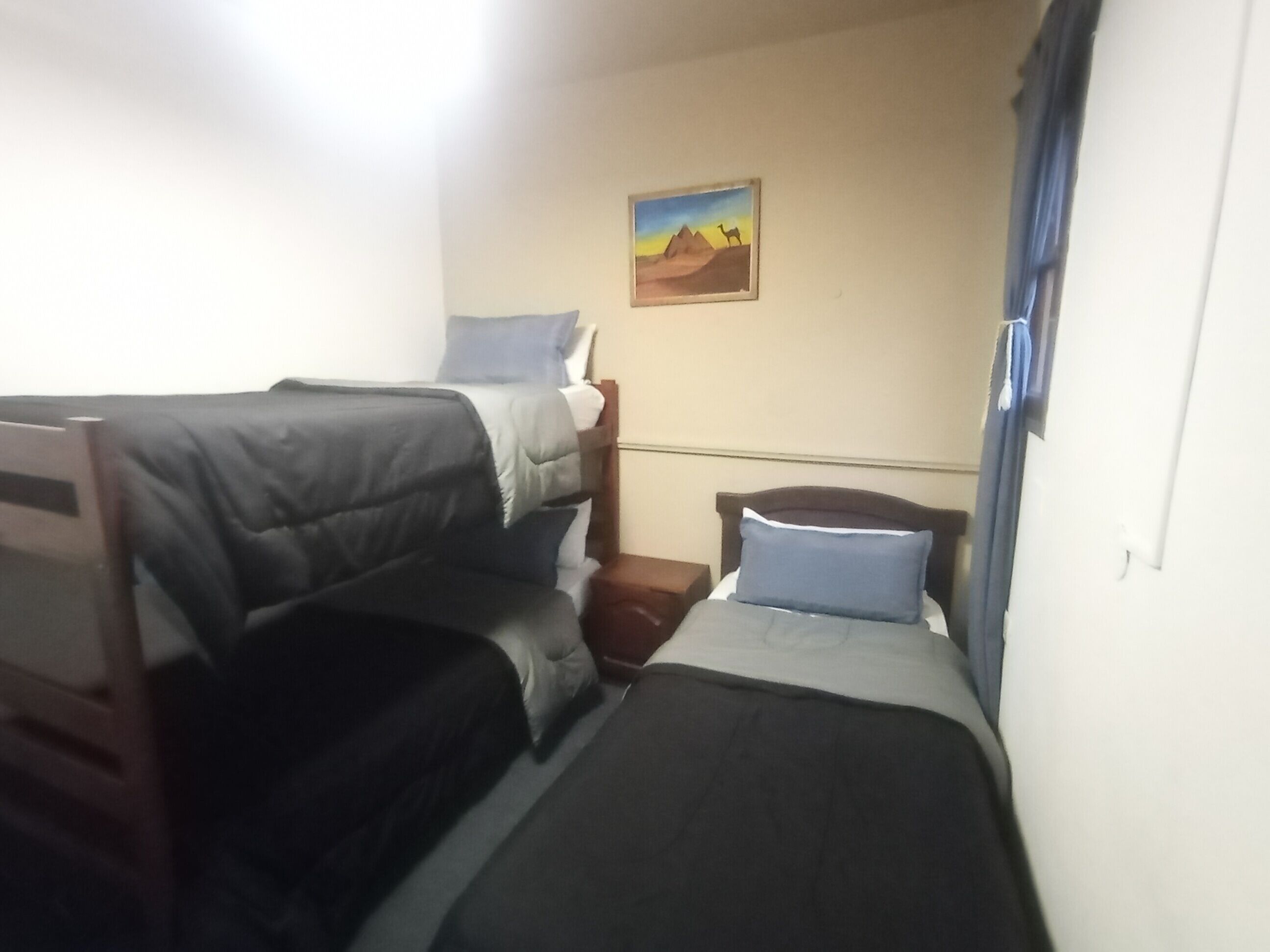 A compact hotel room with two single beds, a small bedside table, a nightstand, and a wall-mounted painting.