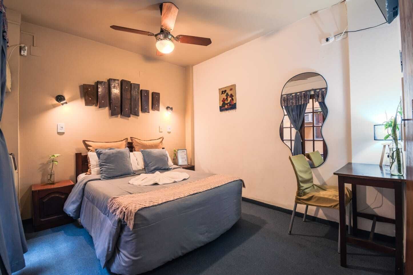 Un dormitorio con cama, mesitas de noche, ventilador de techo, espejo y una pequeña mesa con una planta.