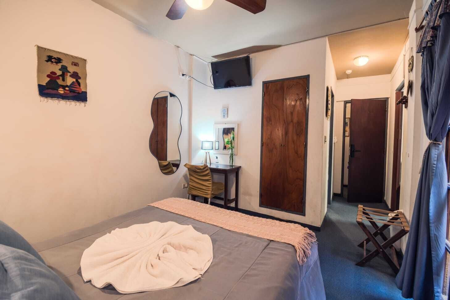 A bedroom with a bed, a desk, a mirror, a TV, and a door.
