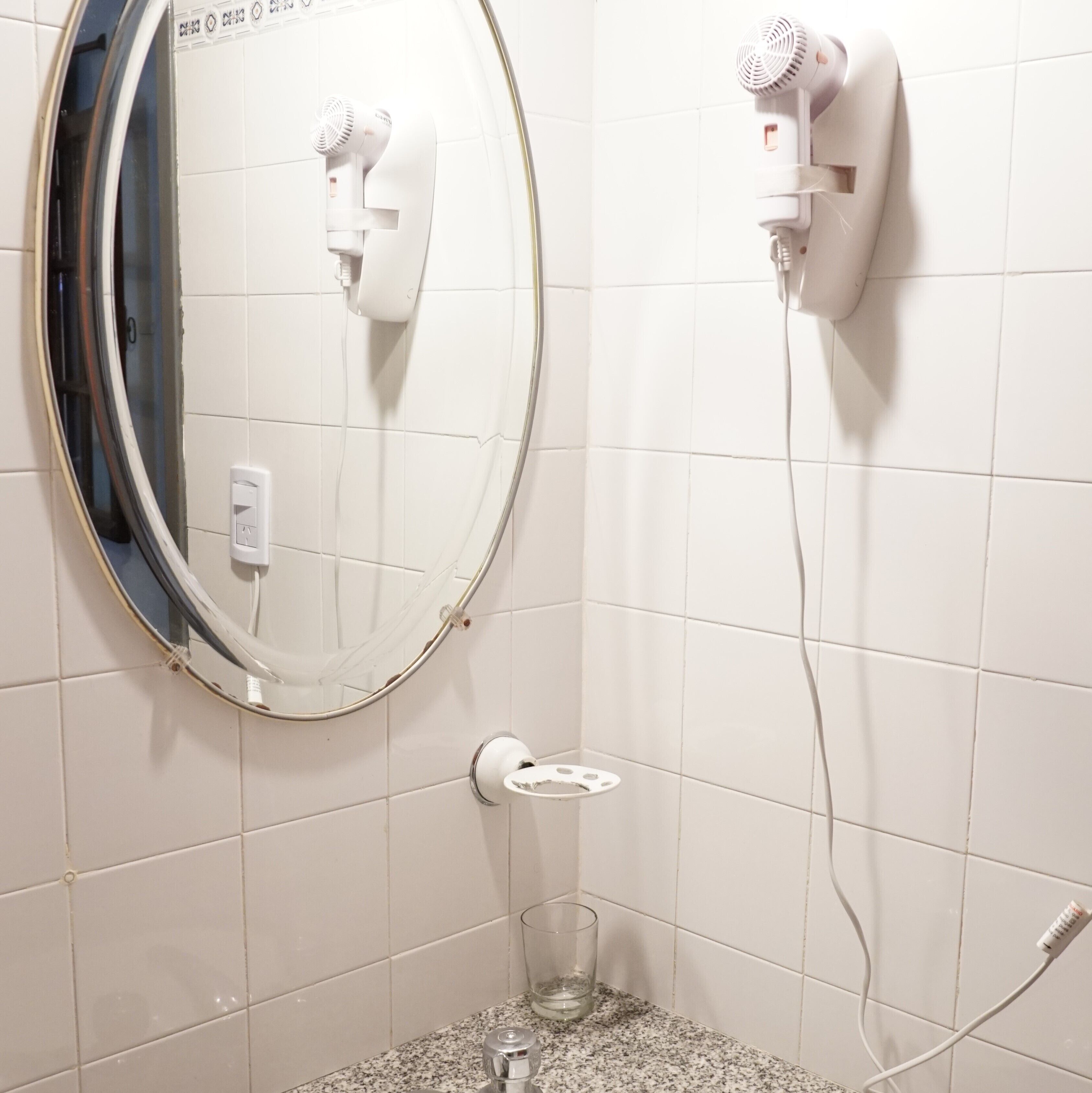 A bathroom with white tiled walls, a round mirror, two hair dryers mounted on the wall, a small glass of water, and a towel rack.