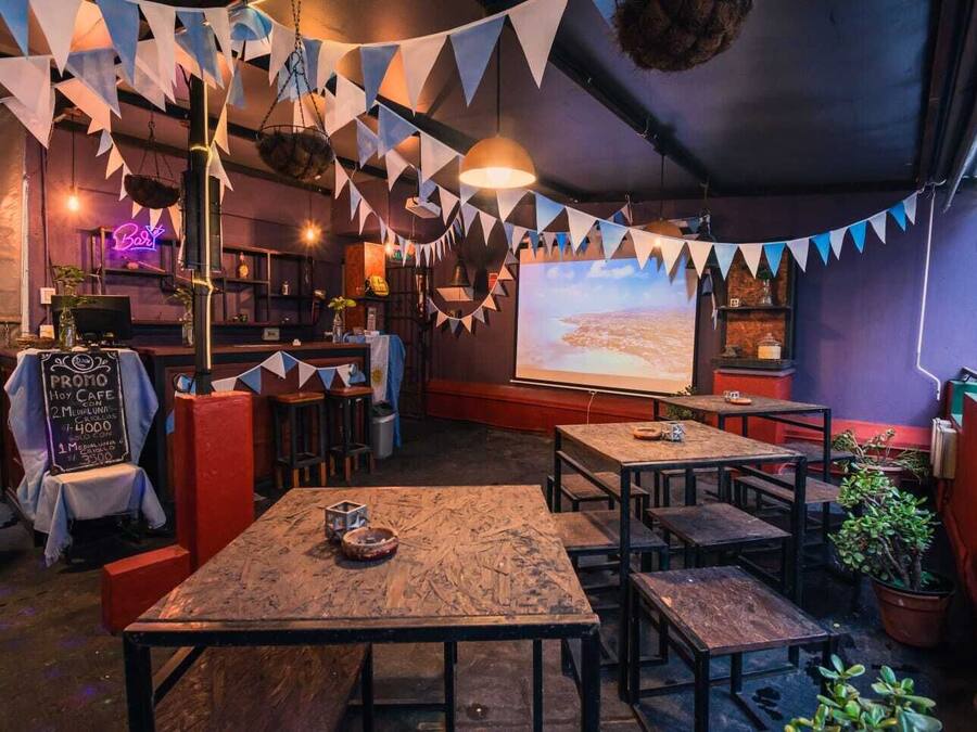 A cozy cafe with wooden tables and chairs, a neon sign, and a large screen displaying a landscape.