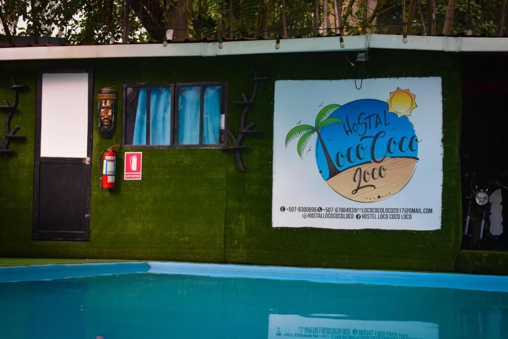 A wall with a sign for 'Hostal Coco Coco' featuring a palm tree and sun, a window with blue curtains, a fire extinguisher, and a door.