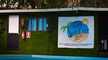 A wall with a sign for 'Hostal Coco Coco' featuring a palm tree and sun, a window with blue curtains, a fire extinguisher, and a door.