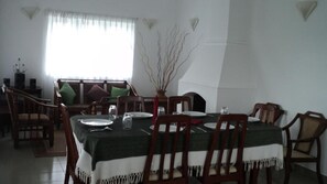 Une salle à manger avec une table dressée pour quatre personnes, entourée de chaises en bois. Un vase contenant des branches est posé sur la table, et on aperçoit une fenêtre avec des rideaux légers au fond.