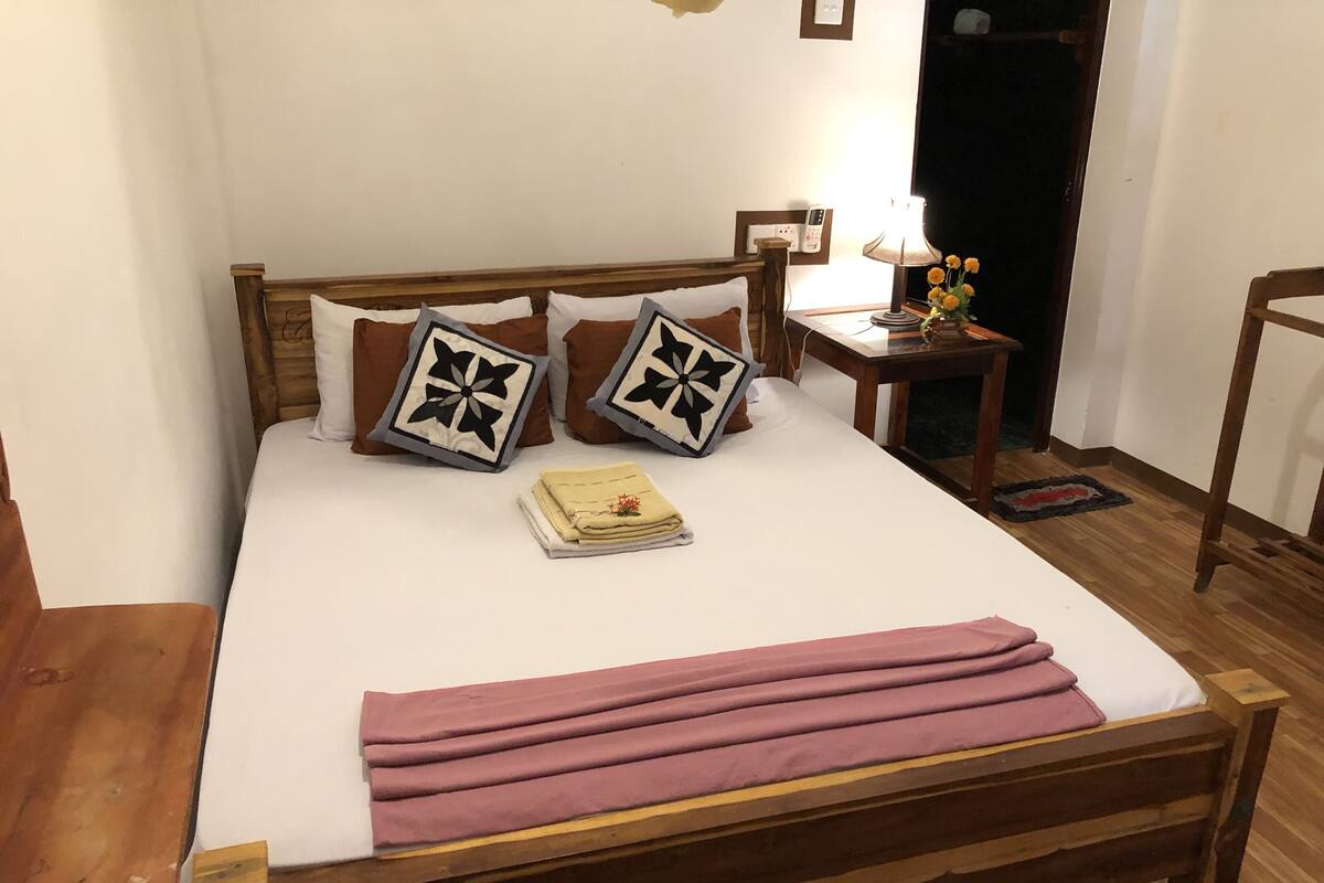 Un lit en bois avec une literie blanche, deux coussins décoratifs, une table de chevet avec une lampe et un petit vase avec des fleurs.