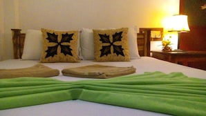 Een net opgemaakt bed met een groene deken, twee decoratieve kussens en een nachtkastje met een lamp en een bloemenvaas.