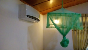 Een kamer met een bed, een houten hoofdbord, een hangend groen klamboes en een aan de muur bevestigde airconditioning.