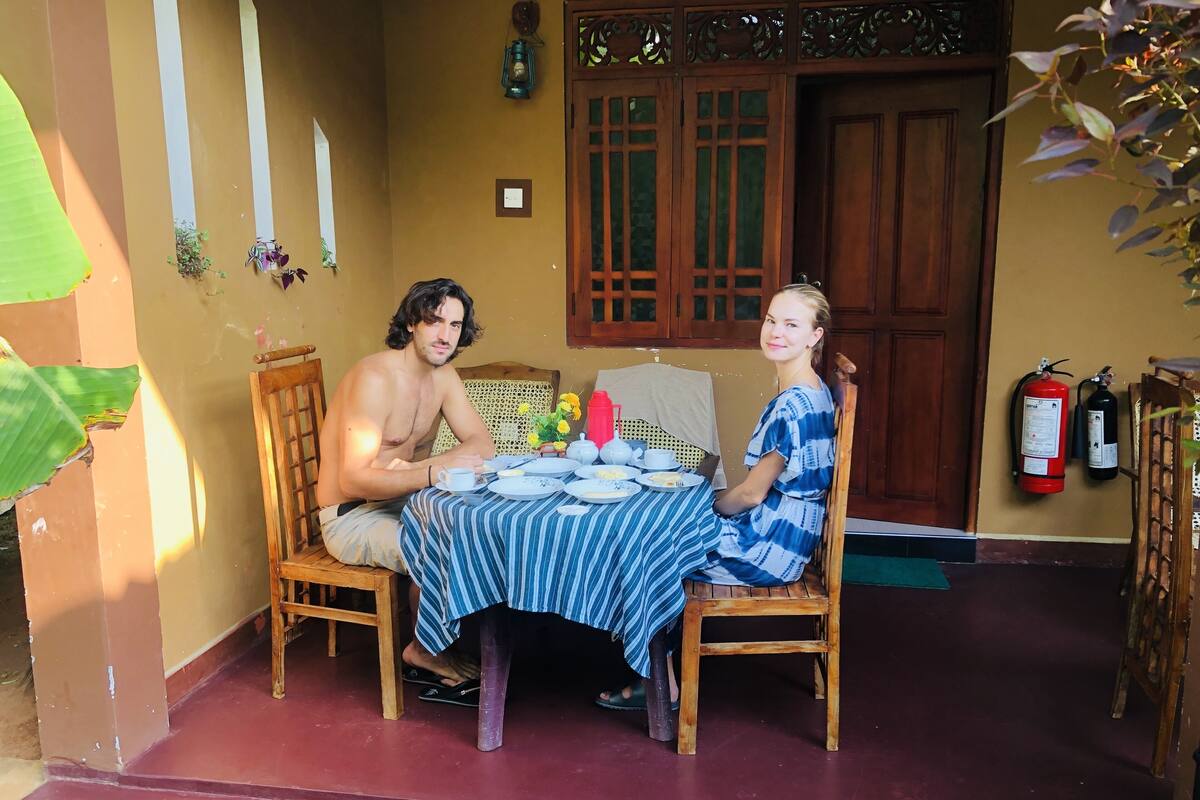 Deux personnes sont assises à une table recouverte d’une nappe à rayures, devant une chaise en bois et une petite table sur laquelle se trouvent des assiettes et des tasses.