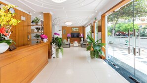 A hotel reception area with a wooden reception desk, a chandelier, and a seating area with sofas and a TV. - Khach San Novena (Ho Chi Minh City)