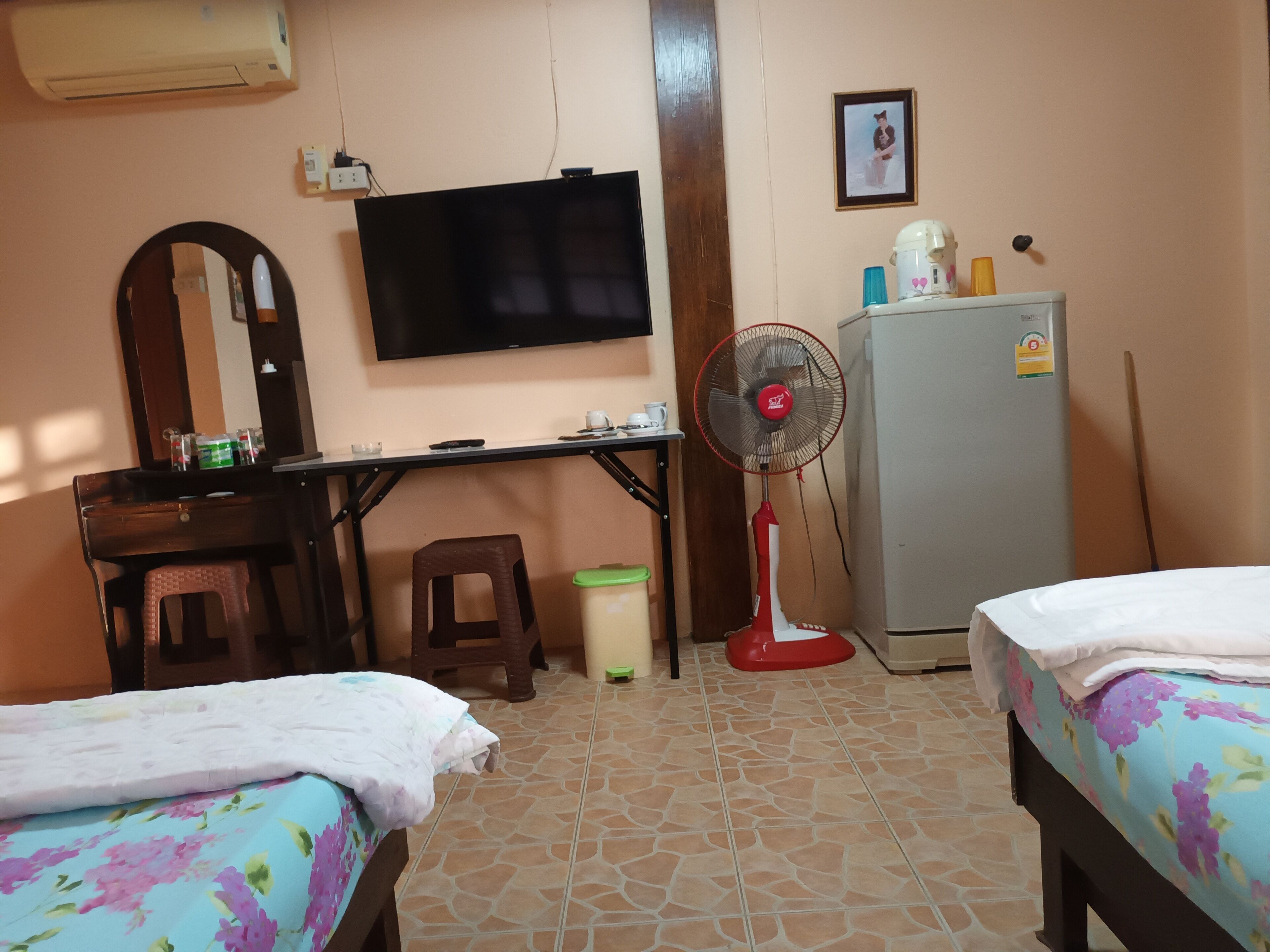 Una habitación con dos camas, un escritorio, un televisor, un ventilador, un refrigerador y un espejo.