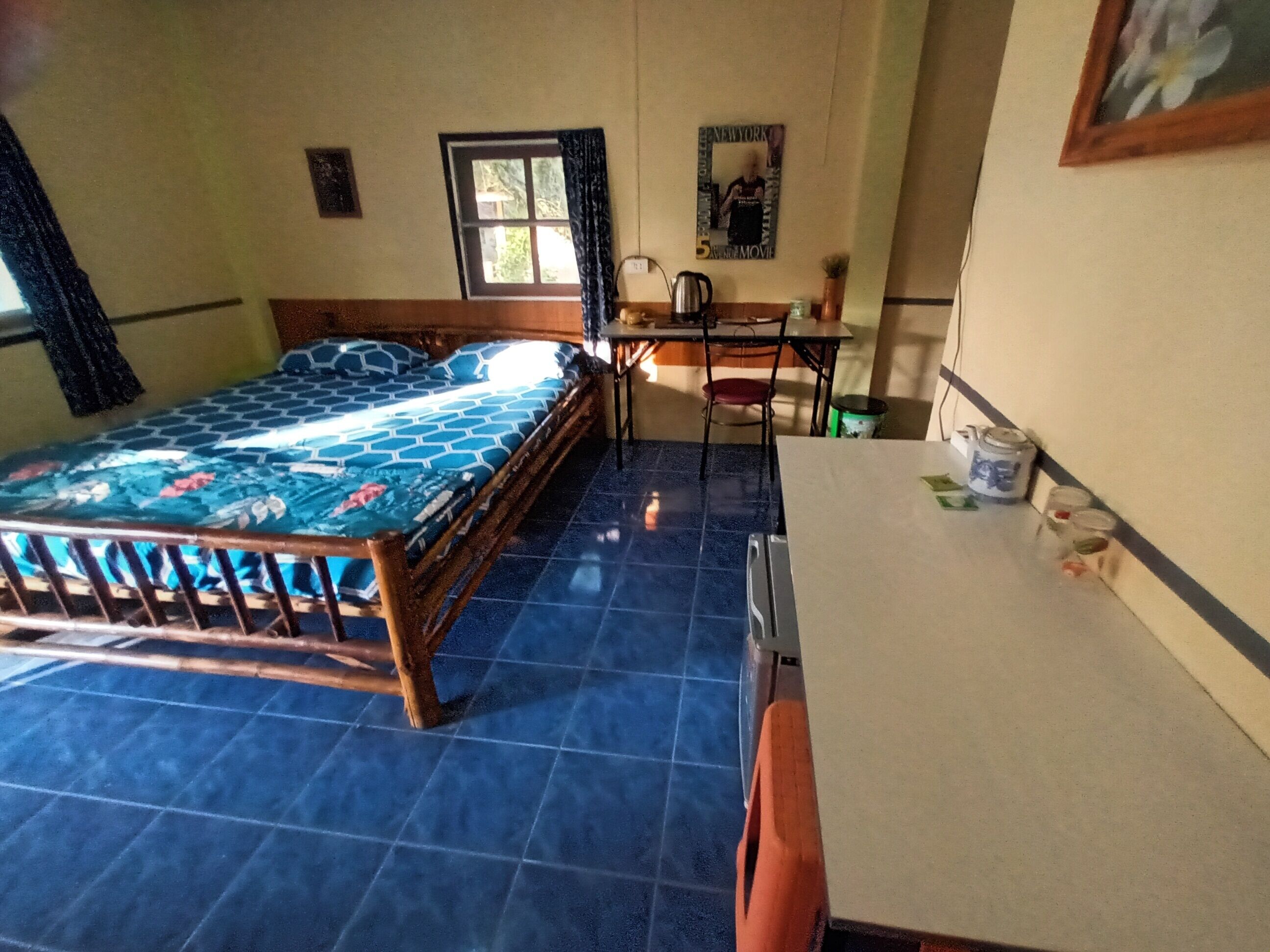 A room with a bed, a desk, and a chair. There is a window with curtains, a picture on the wall, and a small table with a kettle and cups.