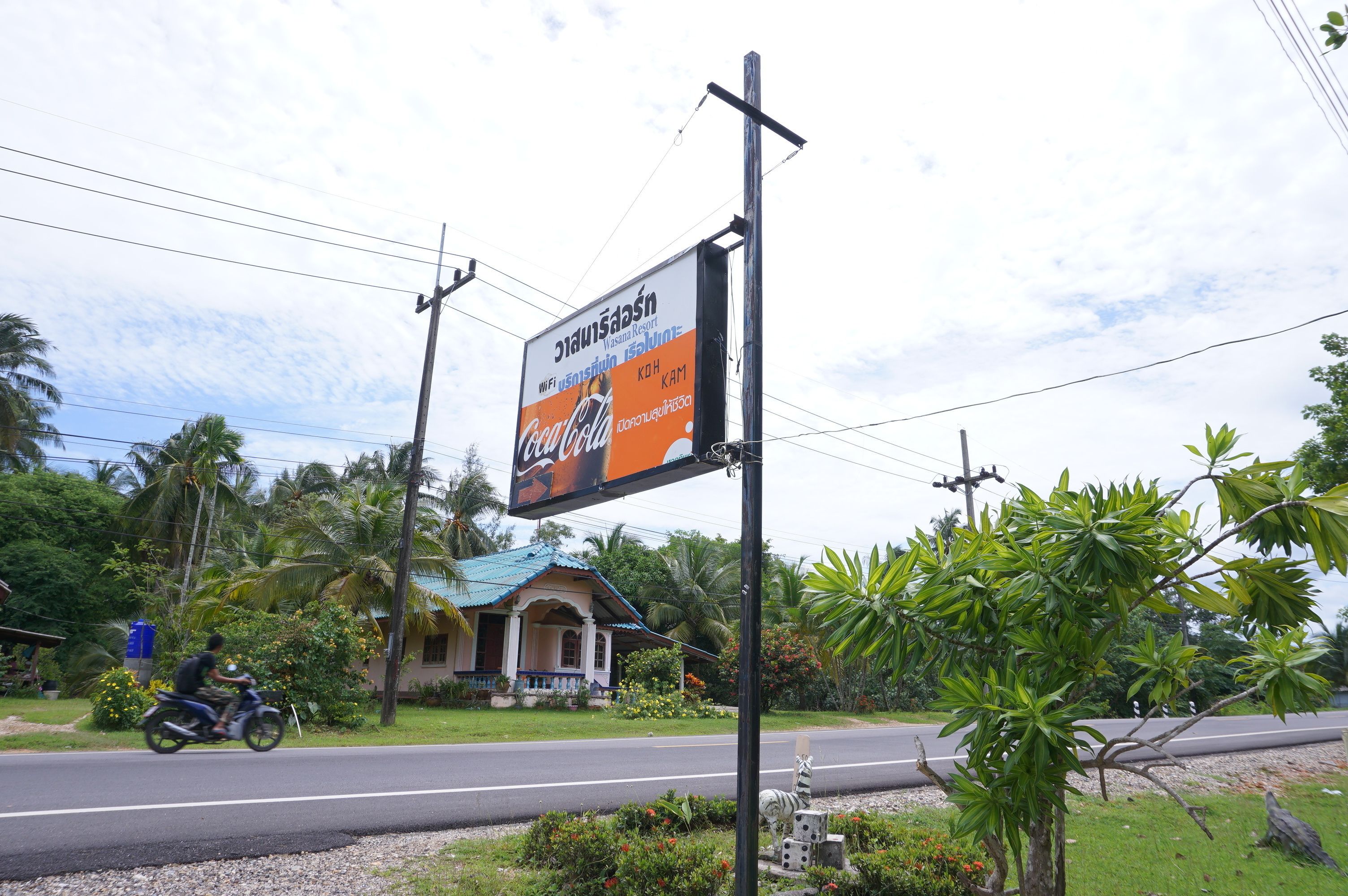 A roadside sign with a Coca-Cola advertisement, a motorbike passing by, and a building with a blue roof in the background.