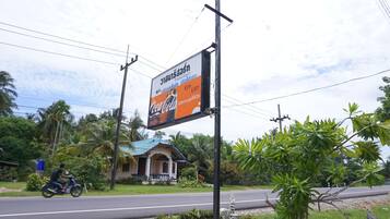 A roadside sign with a Coca-Cola advertisement, a motorbike passing by, and a building with a blue roof in the background.