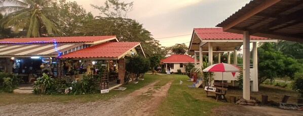 A rustic outdoor setting with a dirt path leading to a building with a red roof. There are small structures with red roofs and a covered area with a bench and umbrella.
