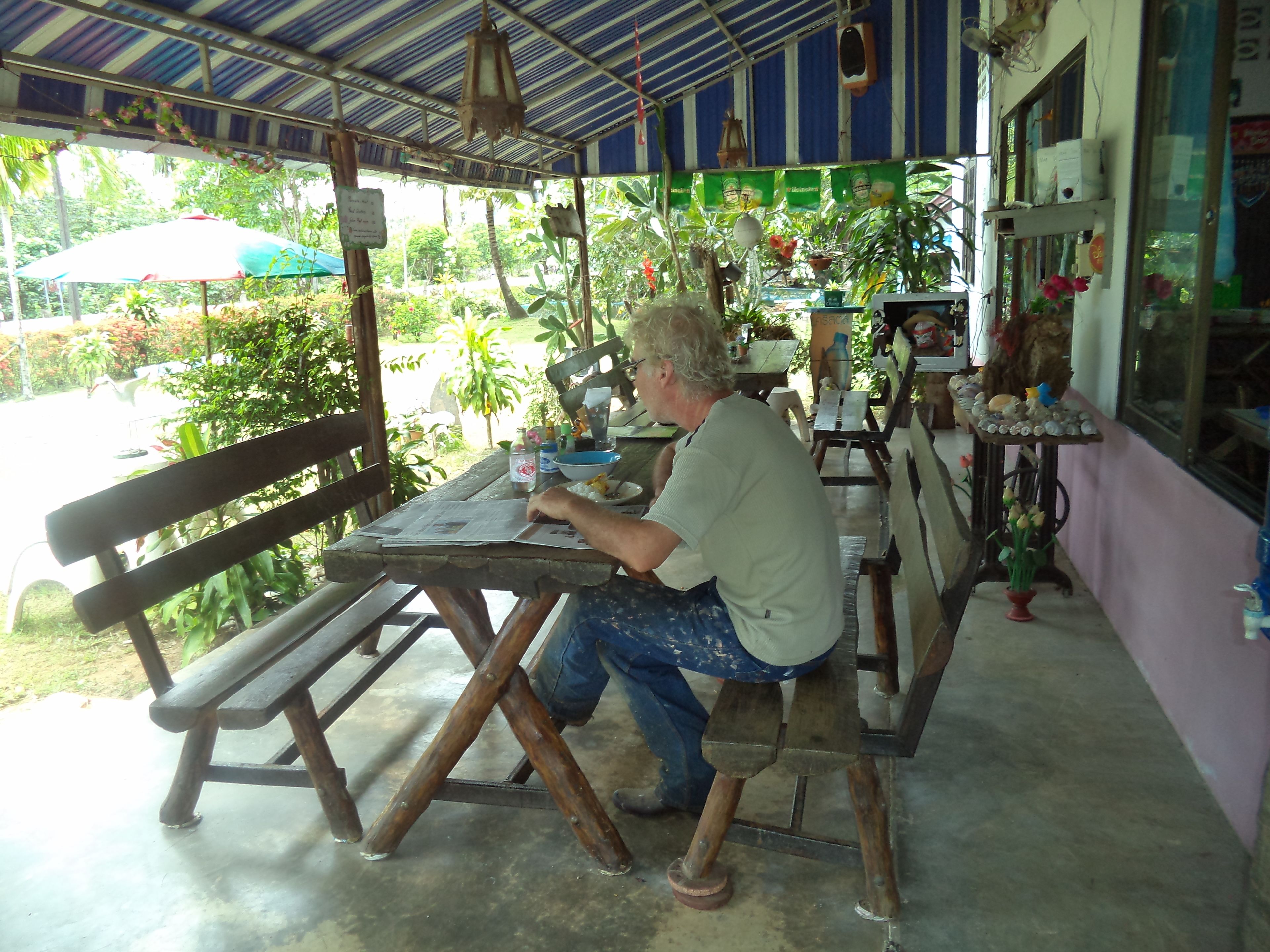 A man sitting at a wooden table under a striped roof, with a bench and a table with a bowl and a bottle on it.