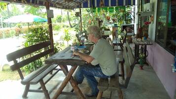 A man sitting at a wooden table under a striped roof, with a bench and a table with a bowl and a bottle on it.