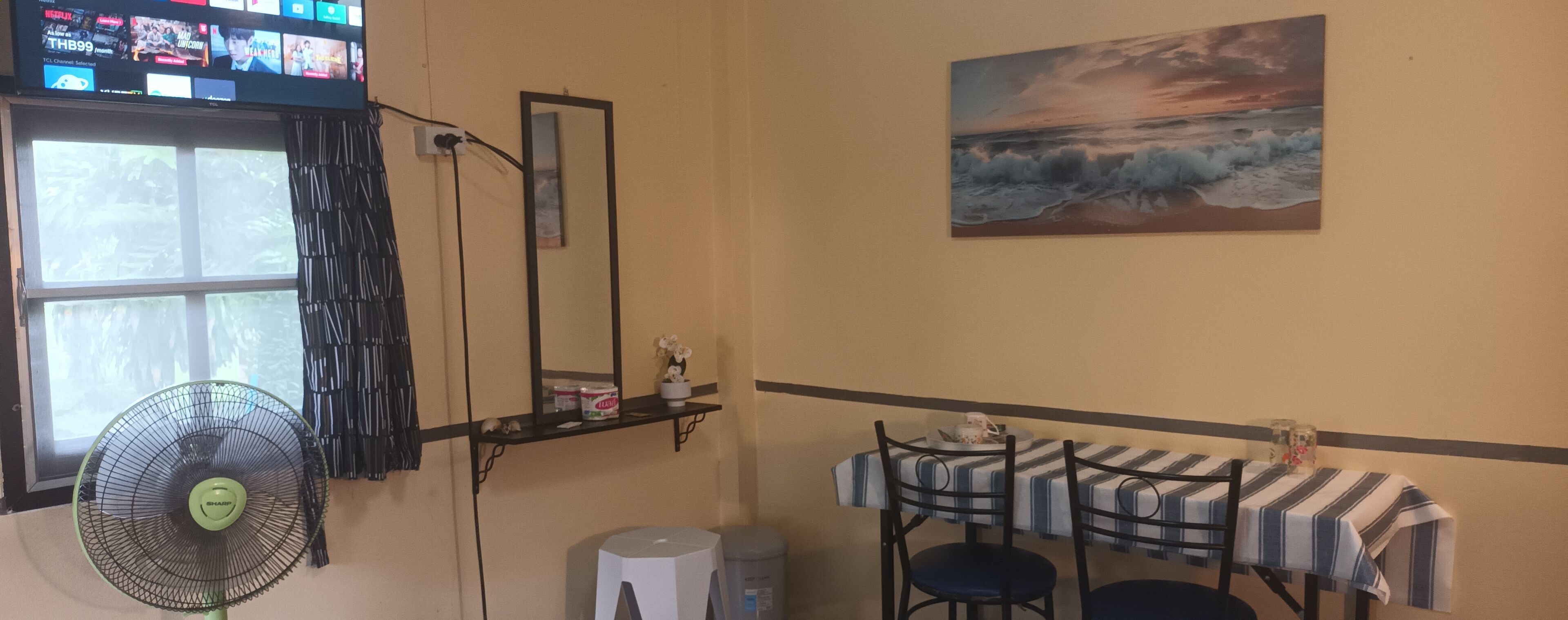 Une pièce équipée d’un téléviseur diffusant différentes chaînes, d’un ventilateur sur pied, d’une table avec des chaises, d’un tableau représentant un coucher de soleil sur l’océan et d’une fenêtre avec des rideaux.